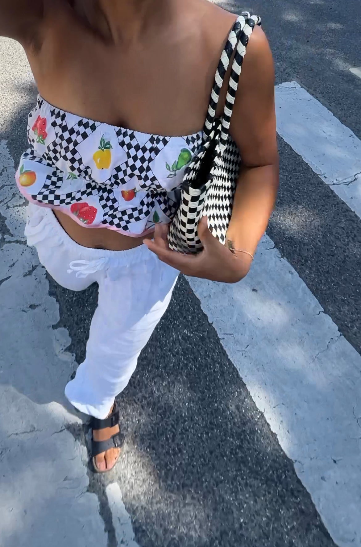 Person wearing a black and white checkered top with fruit pattern and white pants on a street.