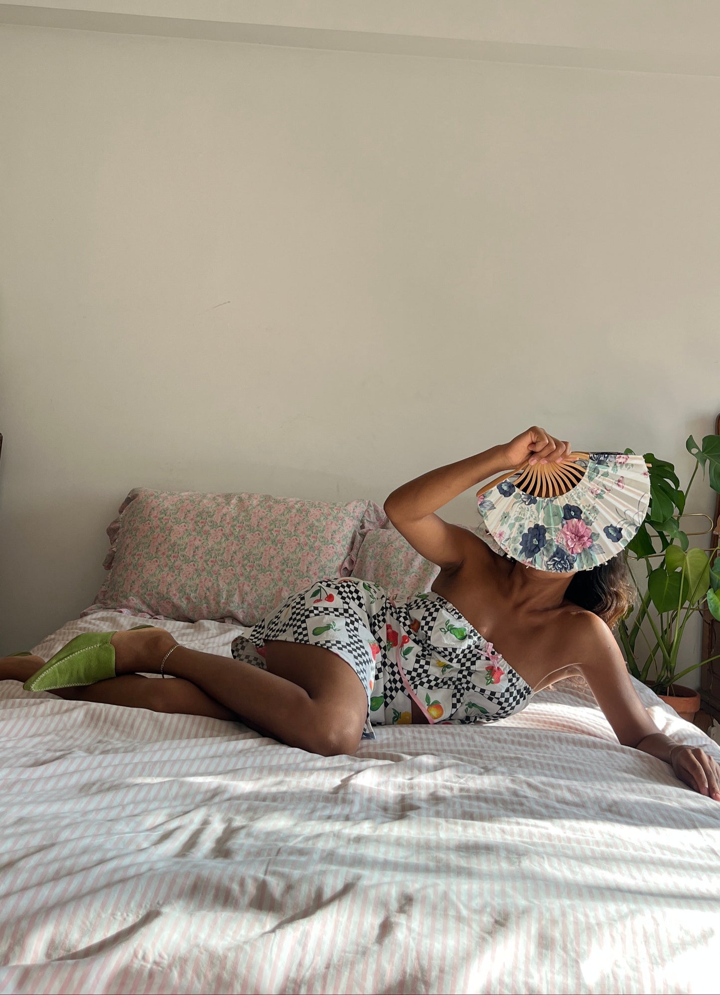 Person sitting on a bed holding a floral fan, wearing a patterned outfit.