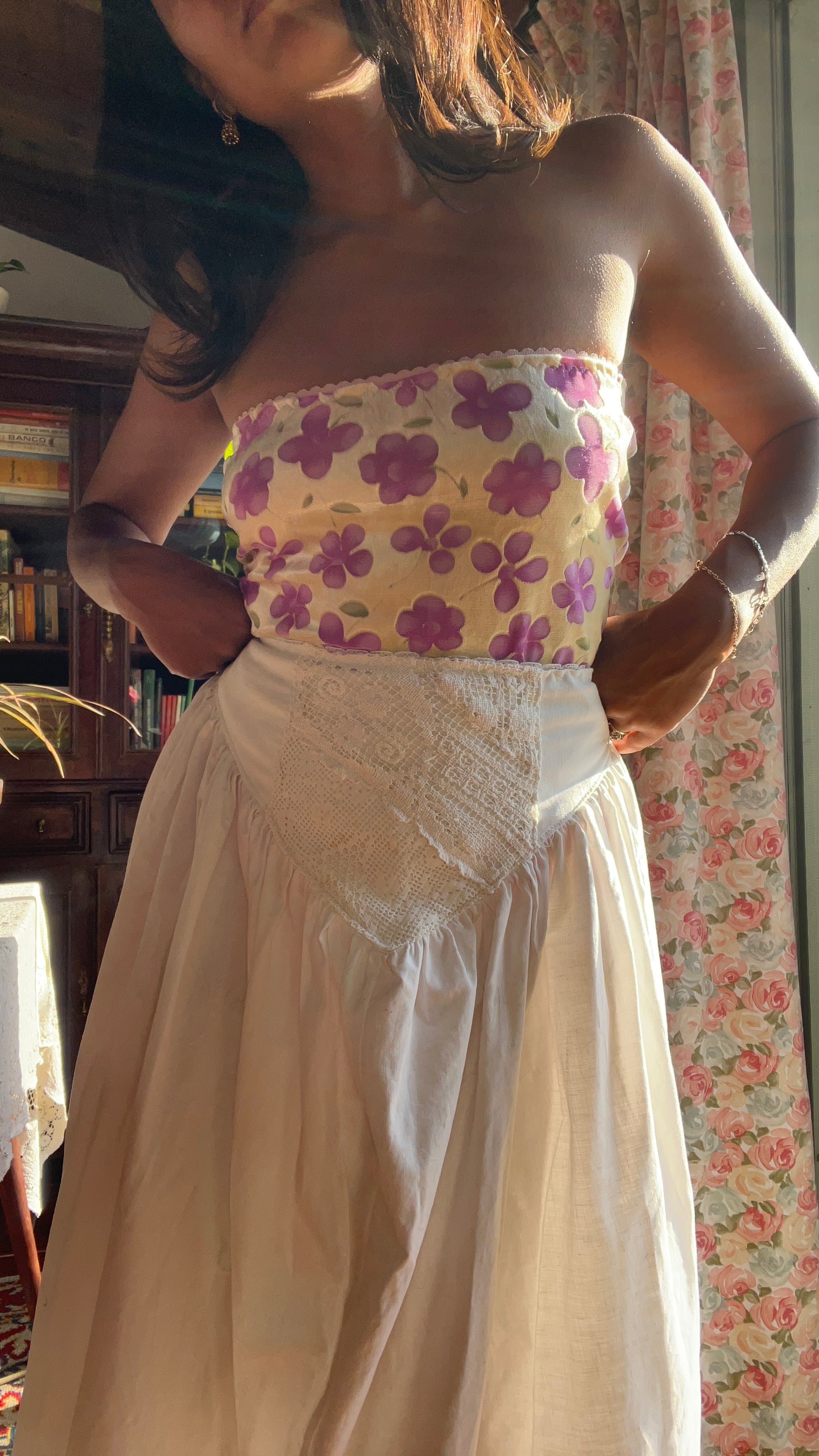 Woman wearing a strapless floral top and white skirt in a room with floral wallpaper. Full outfit hand made by slow fashion brand Yllin. 