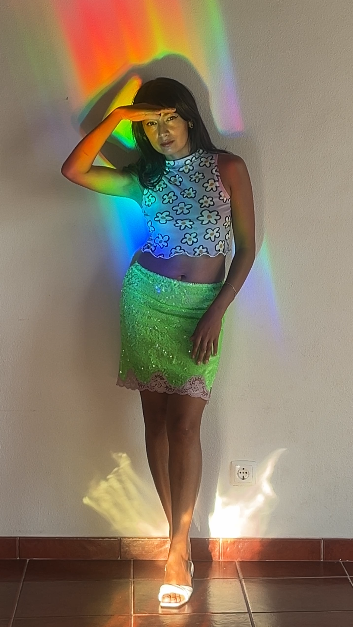 Person standing in front of a wall with a rainbow projection, wearing a colorful top and skirt.