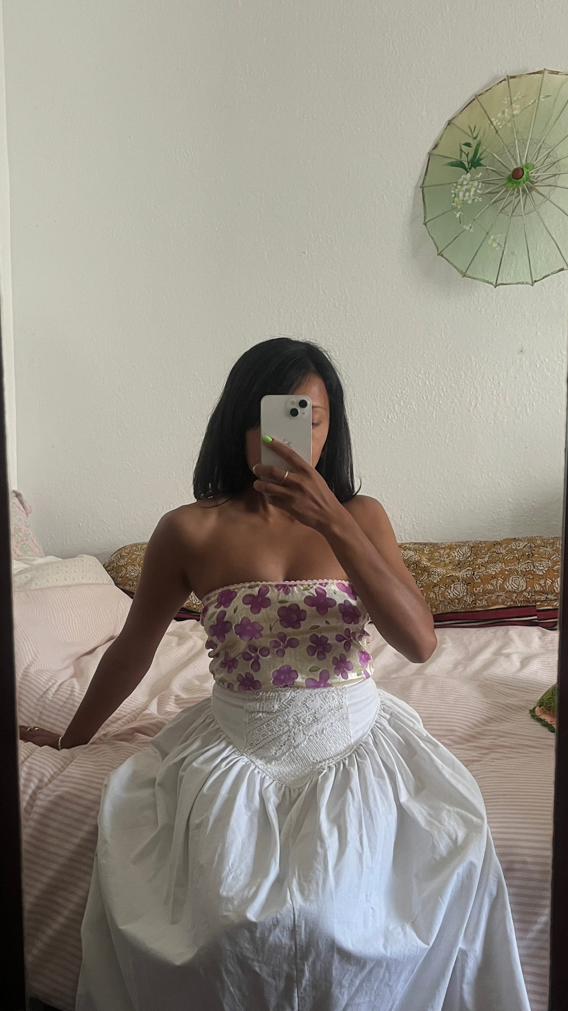Person taking a mirror selfie wearing a floral top and white skirt in a bedroom. Full outfit hand made by slow fashion brand Yllin