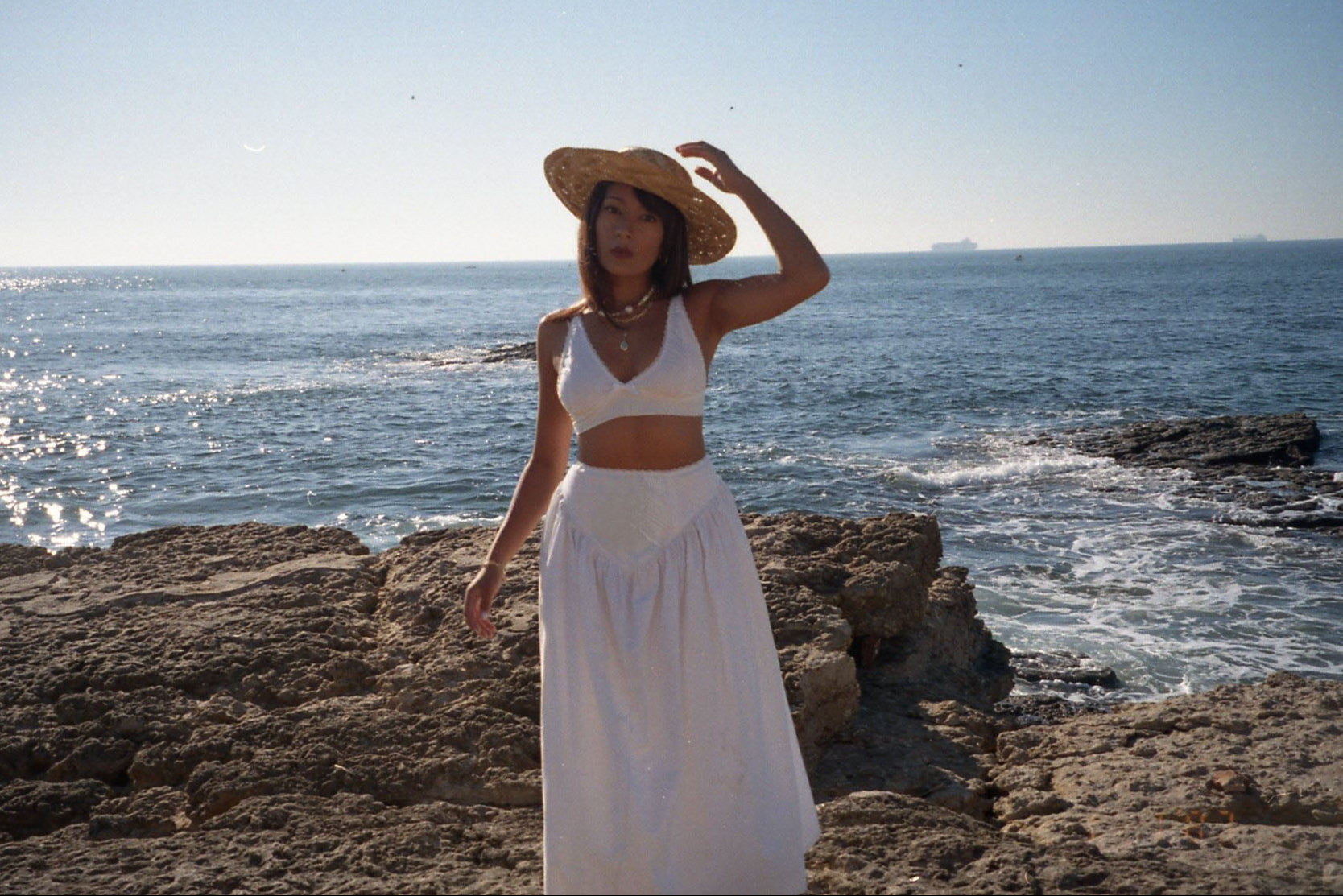 Woman in a white dress and straw hat standing on rocky beach with ocean in background